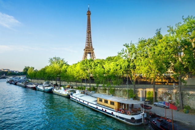 The Eiffel Tower in Paris overlooking the Seine River with houseboats and green trees under a clear blue sky.