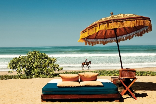 Luxury beach daybed with a traditional Balinese umbrella and a person riding a horse on the shore for a Bali holiday package from Dubai.