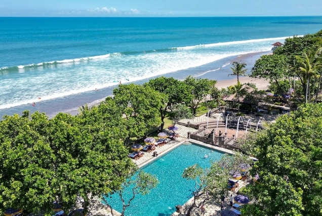 Aerial view of a luxury beachfront resort in Bali with a turquoise swimming pool, tropical palm trees, and ocean waves for Dubai travelers.