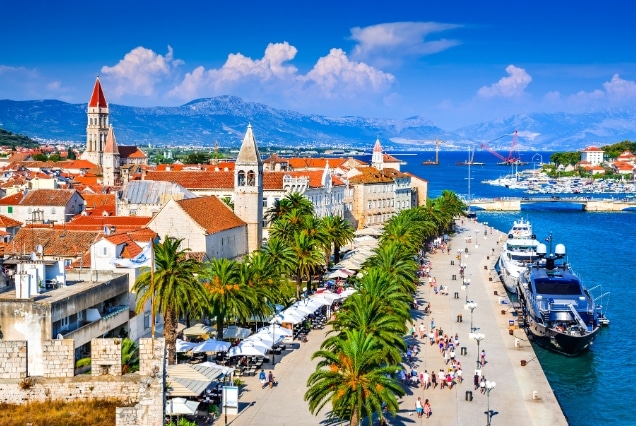 Sunny view of Trogir waterfront promenade with palm trees, yachts, and the Cathedral of St. Lawrence bell tower.