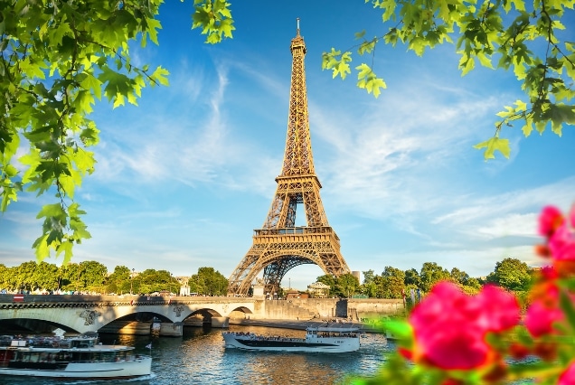 Eiffel Tower framed by green leaves with tour boats on the Seine River under a bright blue sky in Paris.