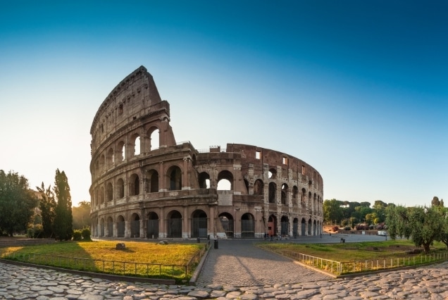 Wide-angle shot of the Roman Colosseum in Italy under a clear blue sky, viewed from a cobblestone path.