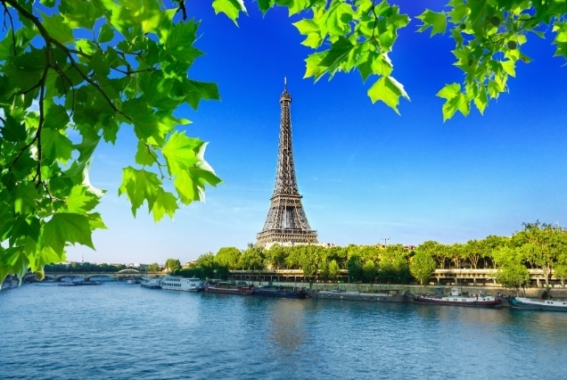 Eiffel Tower in Paris viewed across the Seine River framed by vibrant green summer leaves on a sunny day.