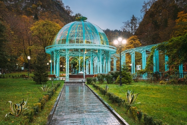 Turquoise glass Mineral Water Pavilion in Borjomi Central Park, Georgia, with autumn trees and a stone path.