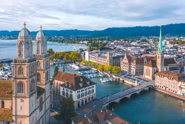 Aerial view of Zurich, Switzerland, featuring the iconic twin towers of Grossmünster church overlooking the Limmat River and Lake Zurich under a soft blue sky.