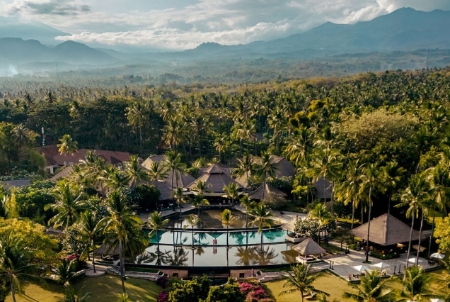 Aerial view of a luxury resort in Lombok with a swimming pool, palm trees, and misty mountains in the background.