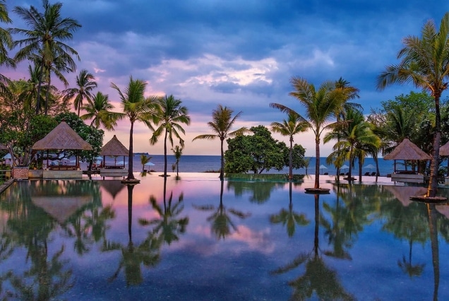 Luxury beachfront infinity pool in Lombok, Indonesia at sunset with palm trees and traditional gazebos.