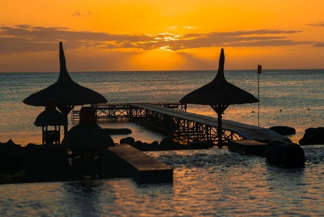 Golden sunset over a Mauritius beach featuring a wooden pier silhouette and traditional straw umbrellas.