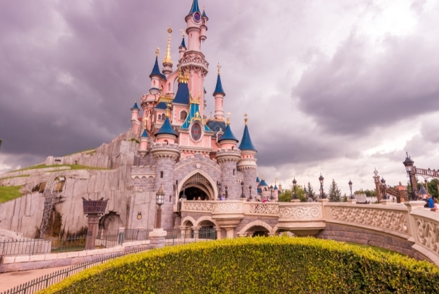 Sleeping Beauty Castle at Disneyland Paris with a stone bridge and dramatic cloudy sky in the background.