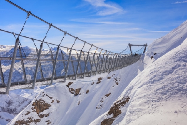 TITLIS Cliff Walk suspension bridge in the Swiss Alps near Engelberg with snow-covered peaks and a clear blue sky.