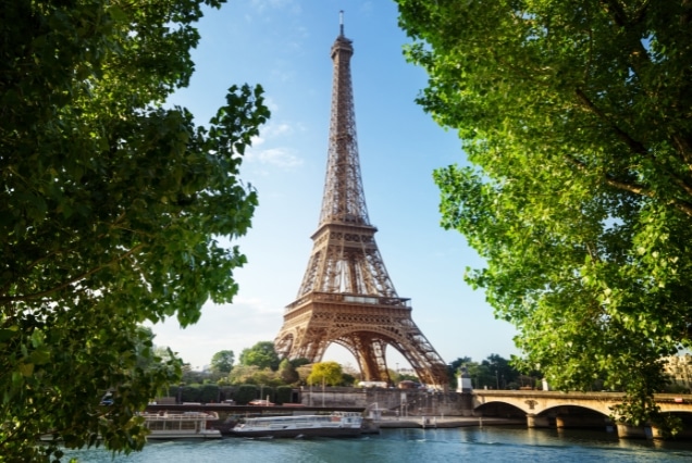 The Eiffel Tower framed by green trees overlooking the Seine River and a stone bridge in Paris, France.