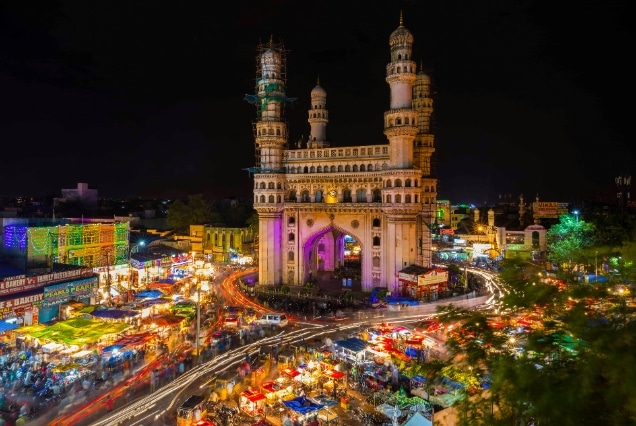 Night view of the illuminated Charminar monument in Hyderabad surrounded by a bustling market and colorful street lights.