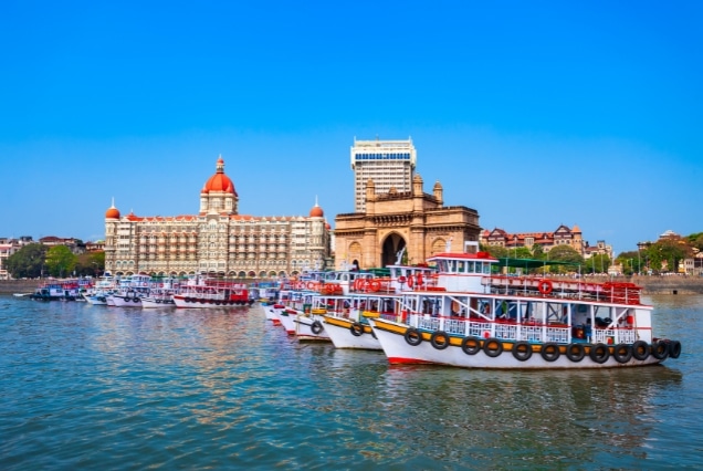 Gateway of India and Taj Mahal Palace Hotel with colorful boats in Mumbai harbor under a clear blue sky.