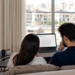 A couple in a UAE apartment looking at a laptop calendar screen trying to book a Schengen visa appointment, with passports nearby.