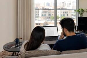 A couple in a UAE apartment looking at a laptop calendar screen trying to book a Schengen visa appointment, with passports nearby.