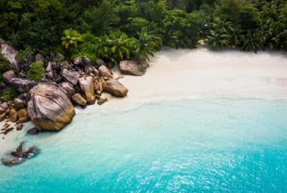 Aerial view of a tropical Seychelles beach with turquoise water, white sand, granite boulders, and lush palm trees.